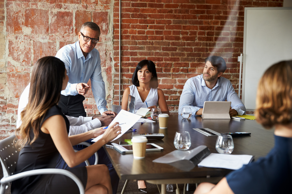 Business professionals sit around a conference table in a brick-walled meeting room while a man stands and leads the discussion.