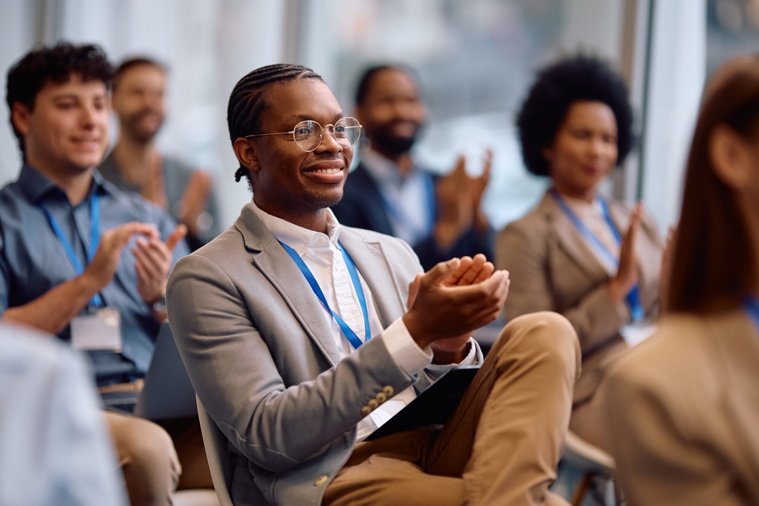 Conference attendees wearing lanyards applaud during a presentation, with a smiling man in glasses seated in the foreground.